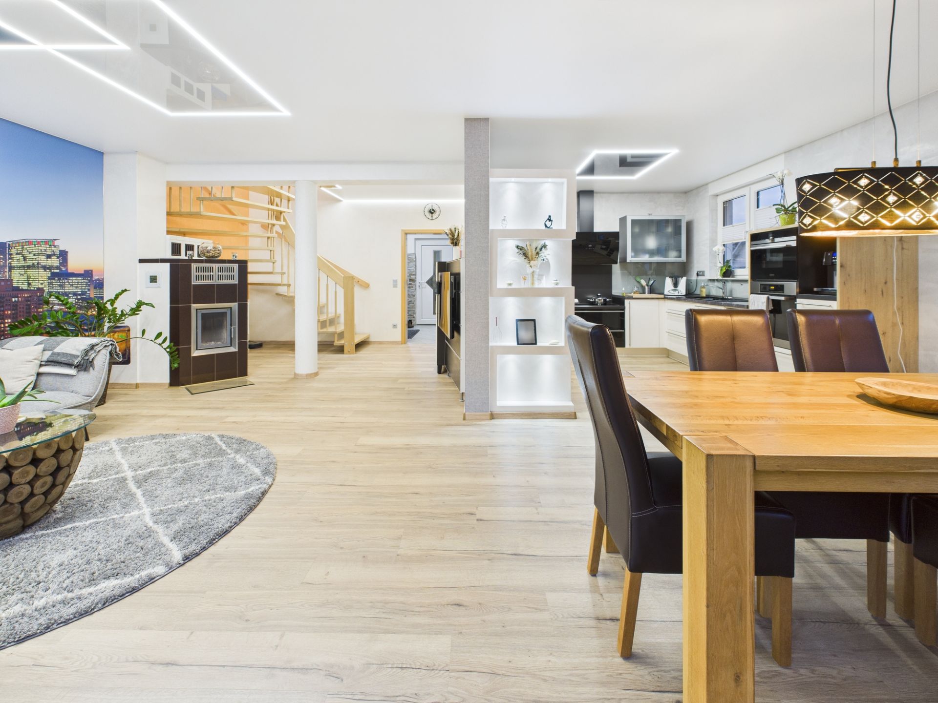 Modernes Wohnzimmer mit Essbereich, offener Küche und Holztreppe im Hintergrund.