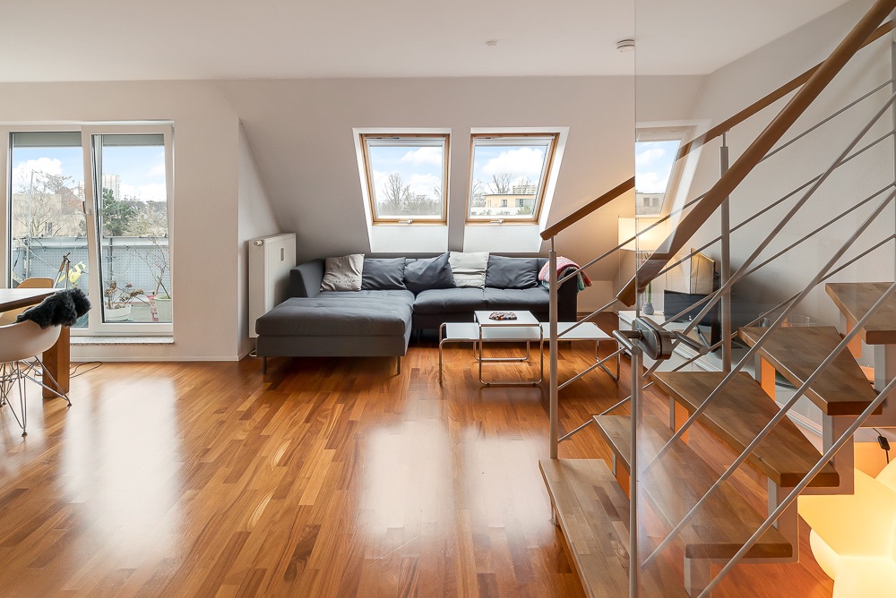Helles Wohnzimmer mit Holzfußboden, Ecksofa, Couchtisch und Treppe, zwei Dachfenster im Hintergrund.