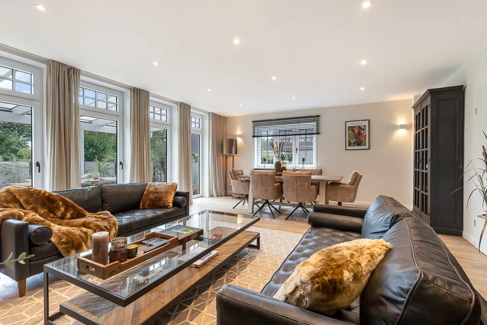 Modernes Wohnzimmer mit zwei Ledersofas, Glastisch und Essbereich vor großen Fenstern.