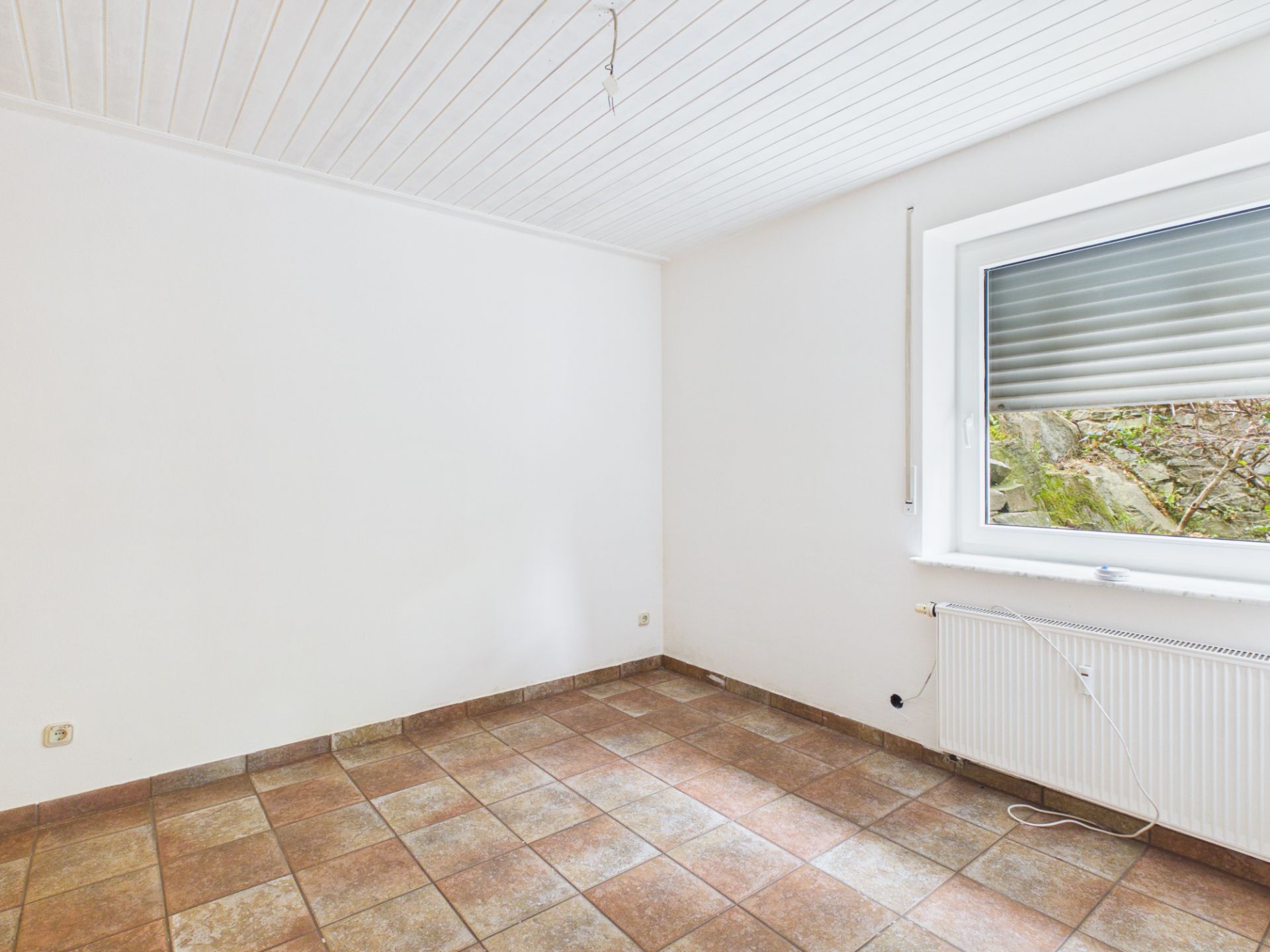 Leerer Raum mit Fliesenboden, weißer Decke und Fenster mit Rollladen.