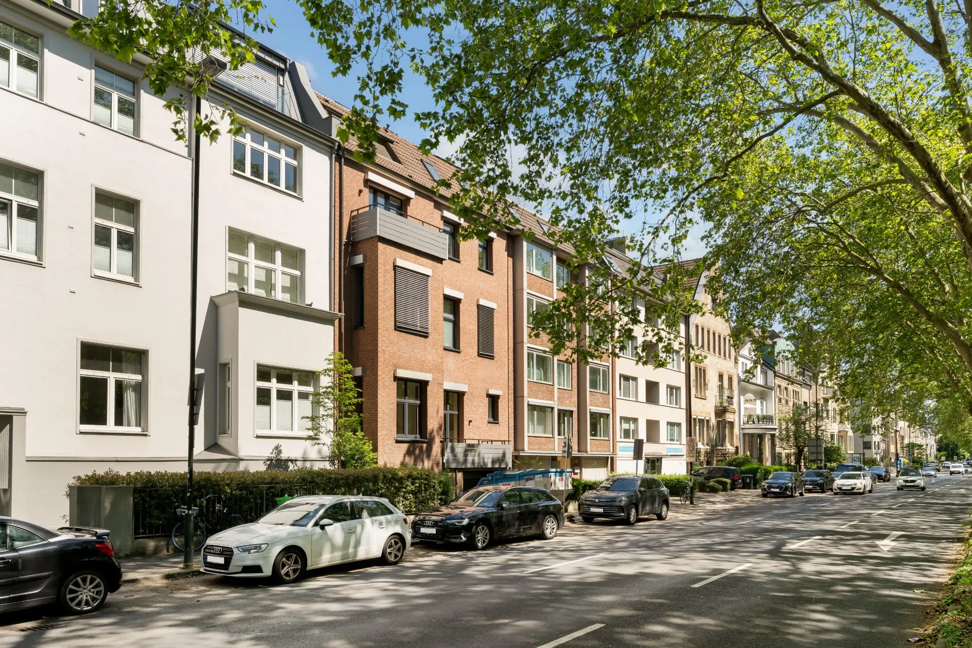 Straße mit geparkten Autos und mehreren Wohnhäusern, Bäume bieten Schatten.