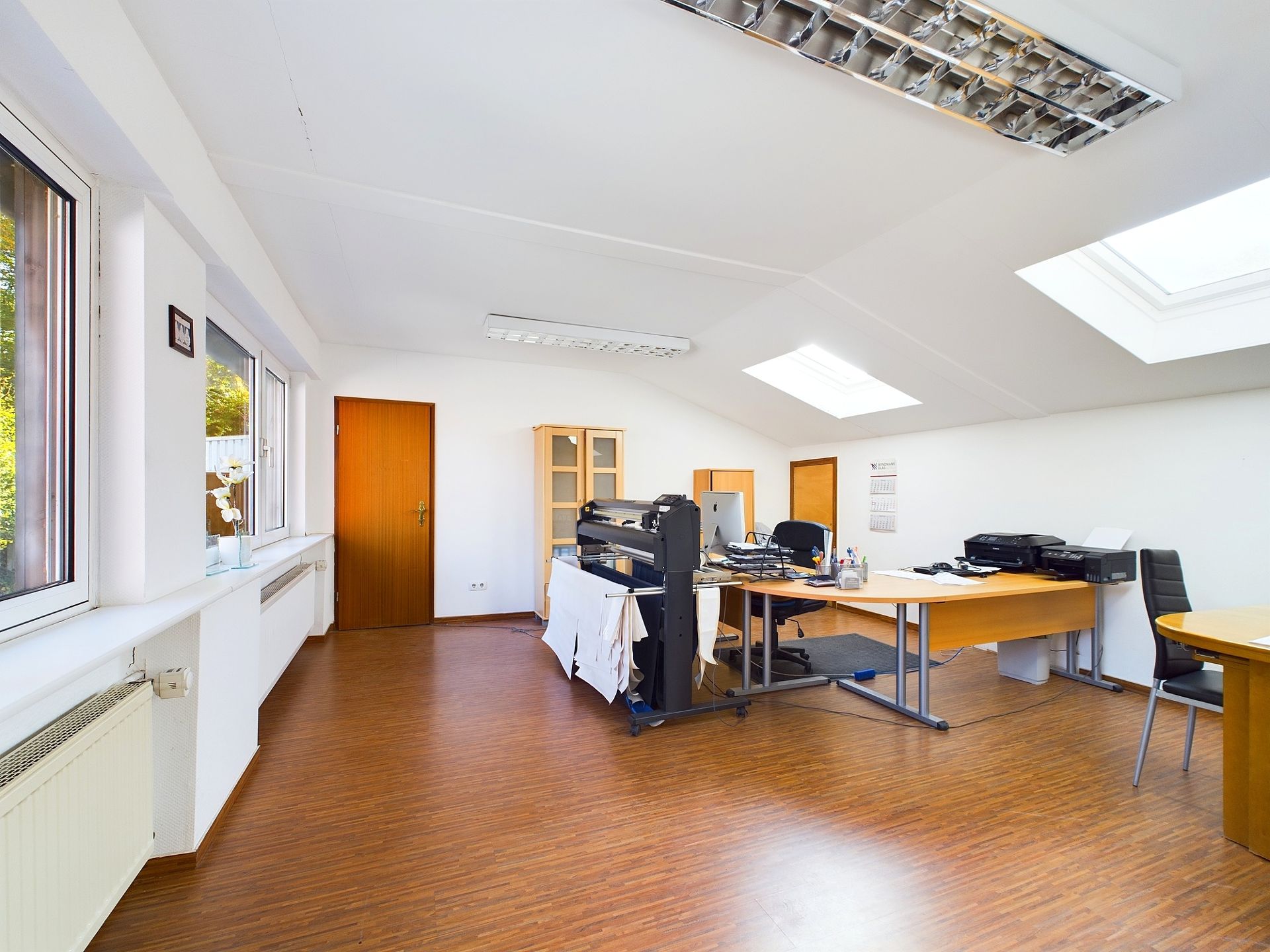 Helles Arbeitszimmer mit Schreibtischen, Stühlen, Druckern und großen Fenstern.