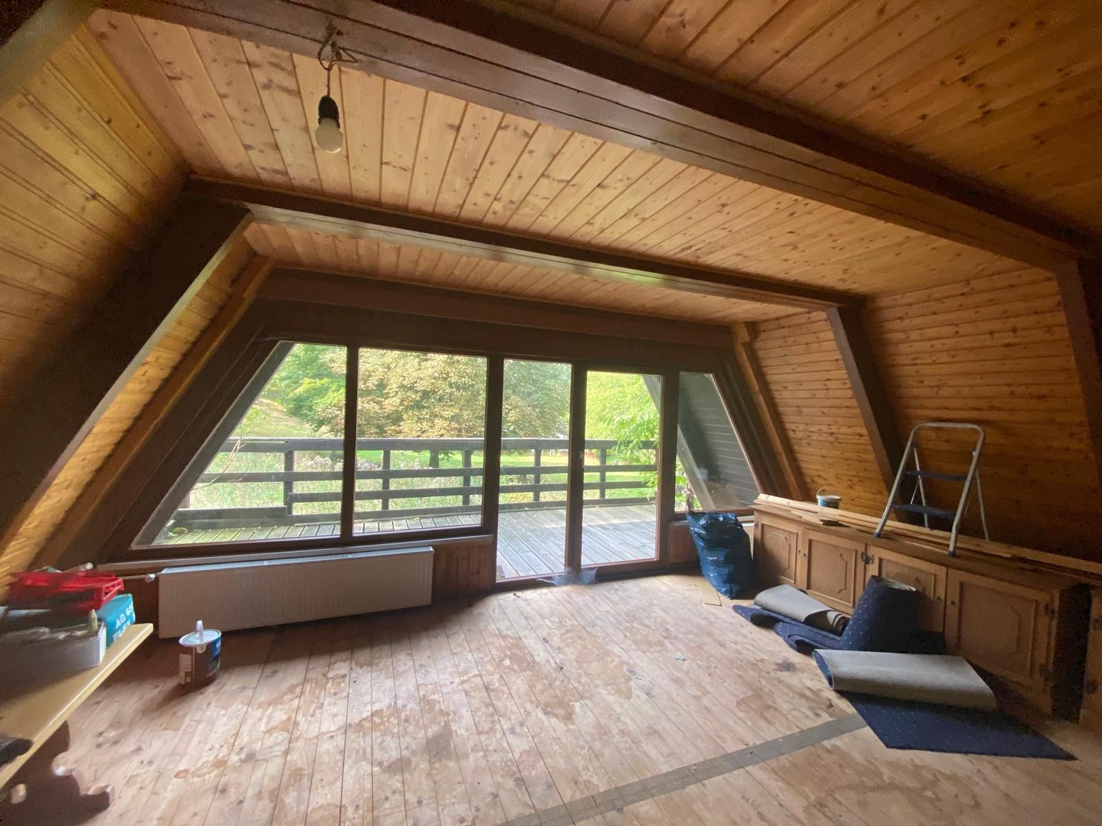 Holzverkleideter Raum mit großen Fenstern und Zugang zu einem Balkon, einige Gegenstände am Boden.