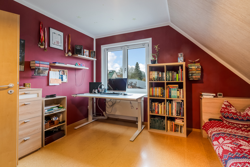 Arbeitszimmer mit Schreibtisch, Regal, Bett und roter Wand unter Dachschräge.