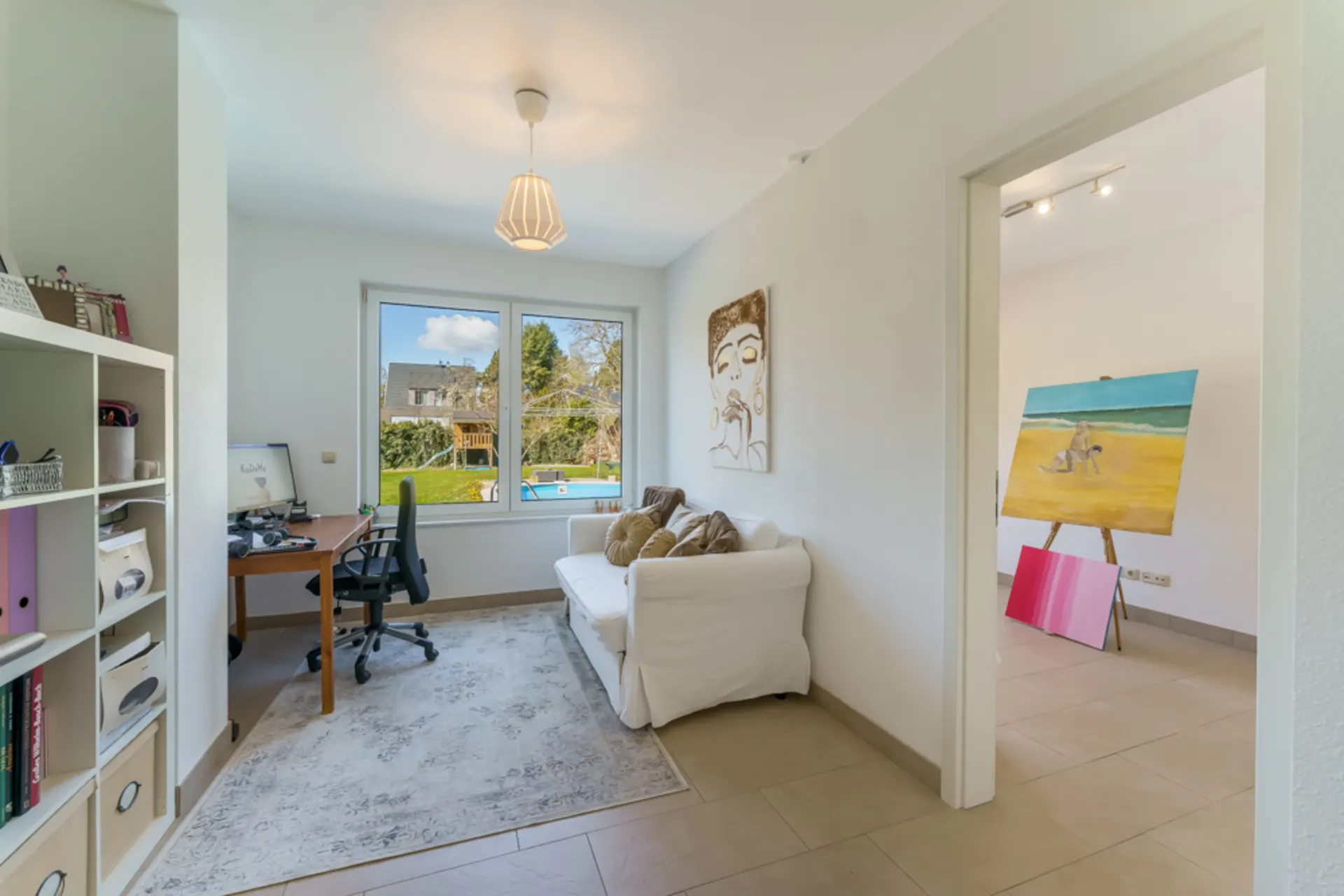 Helles Zimmer mit Sofa, Schreibtisch, Regal und Zugang zu einem weiteren Raum mit Staffelei.