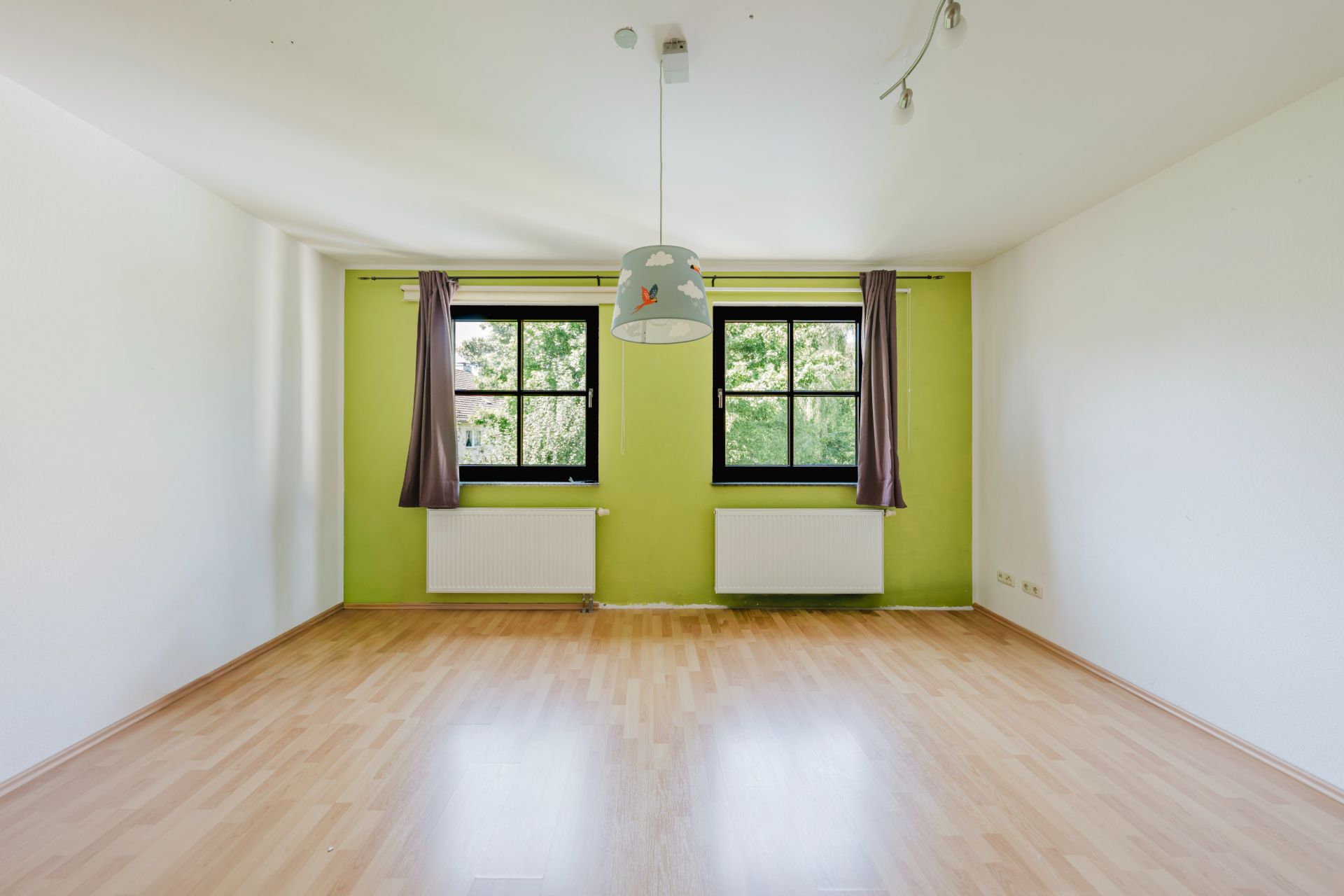 Leerer Raum mit hellem Holzboden, zwei Fenstern, grüner Wand und Deckenlampe.