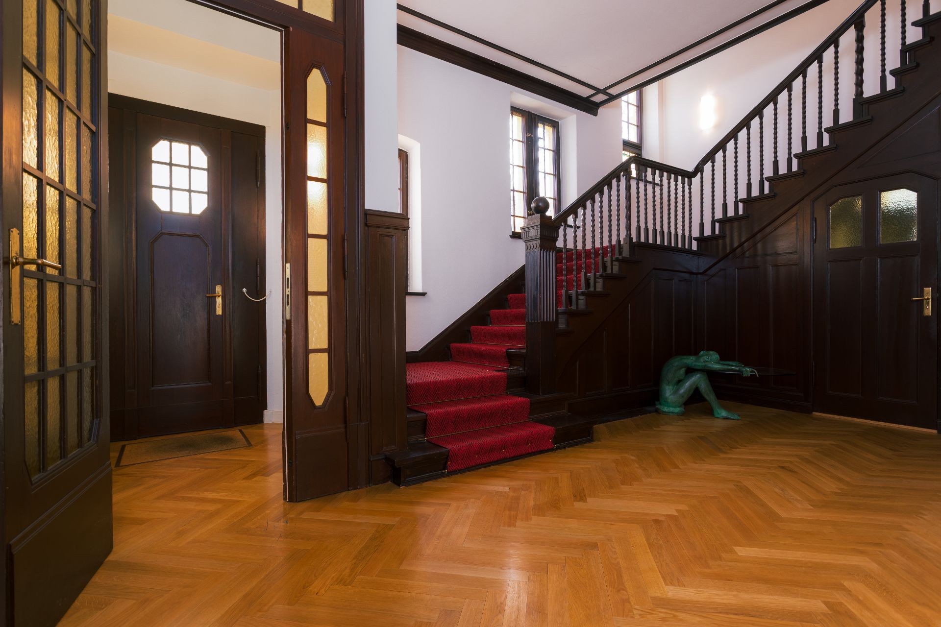 Flur mit Holztreppe, rotem Teppichläufer und grünlicher Skulptur unter der Treppe.