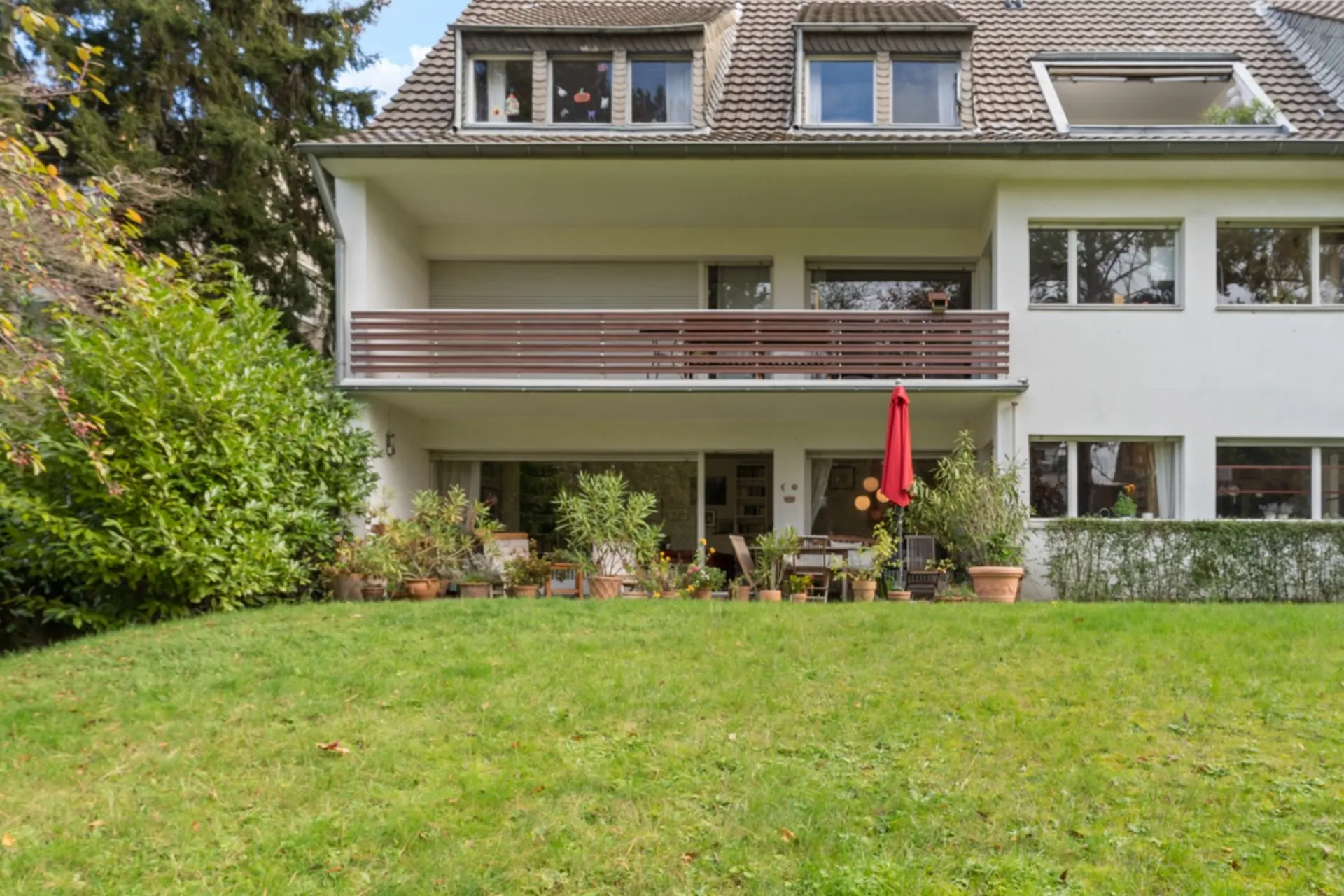Weiße Wohnhausfassade mit Balkon, Terrasse, vielen Pflanzen und rotem Sonnenschirm vor einer Rasenfläche.