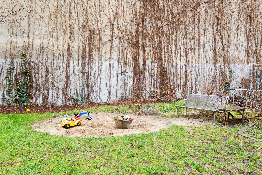 Garten mit Sandkasten, Kinderspielzeug, einer Bank und Fahrrädern an einer Hauswand mit Kletterpflanzen.