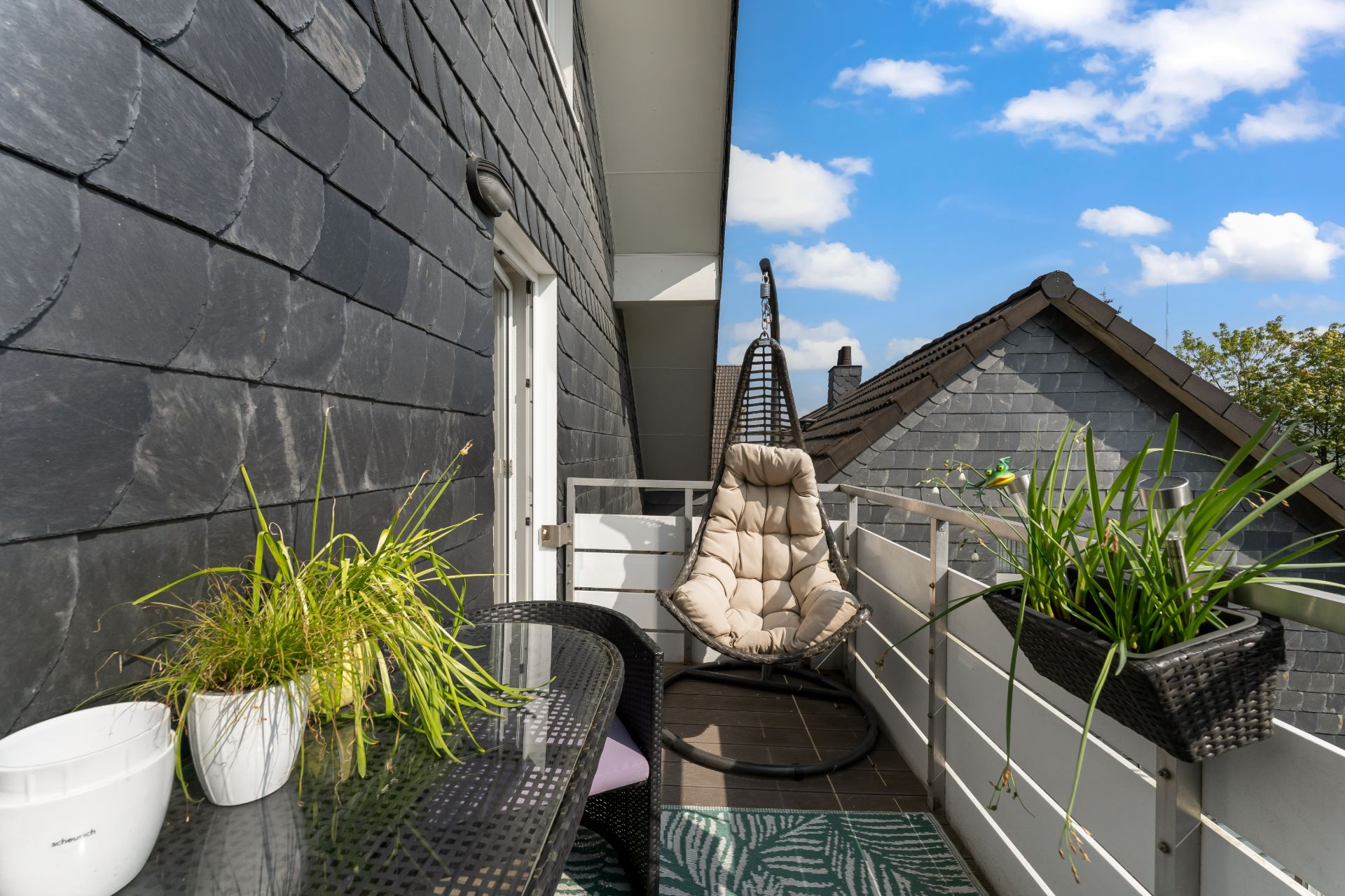 Balkon mit Hängekorb-Stuhl, Tisch, Stühlen und Pflanzen, daneben Schieferwände und Blick auf Hausdächer.