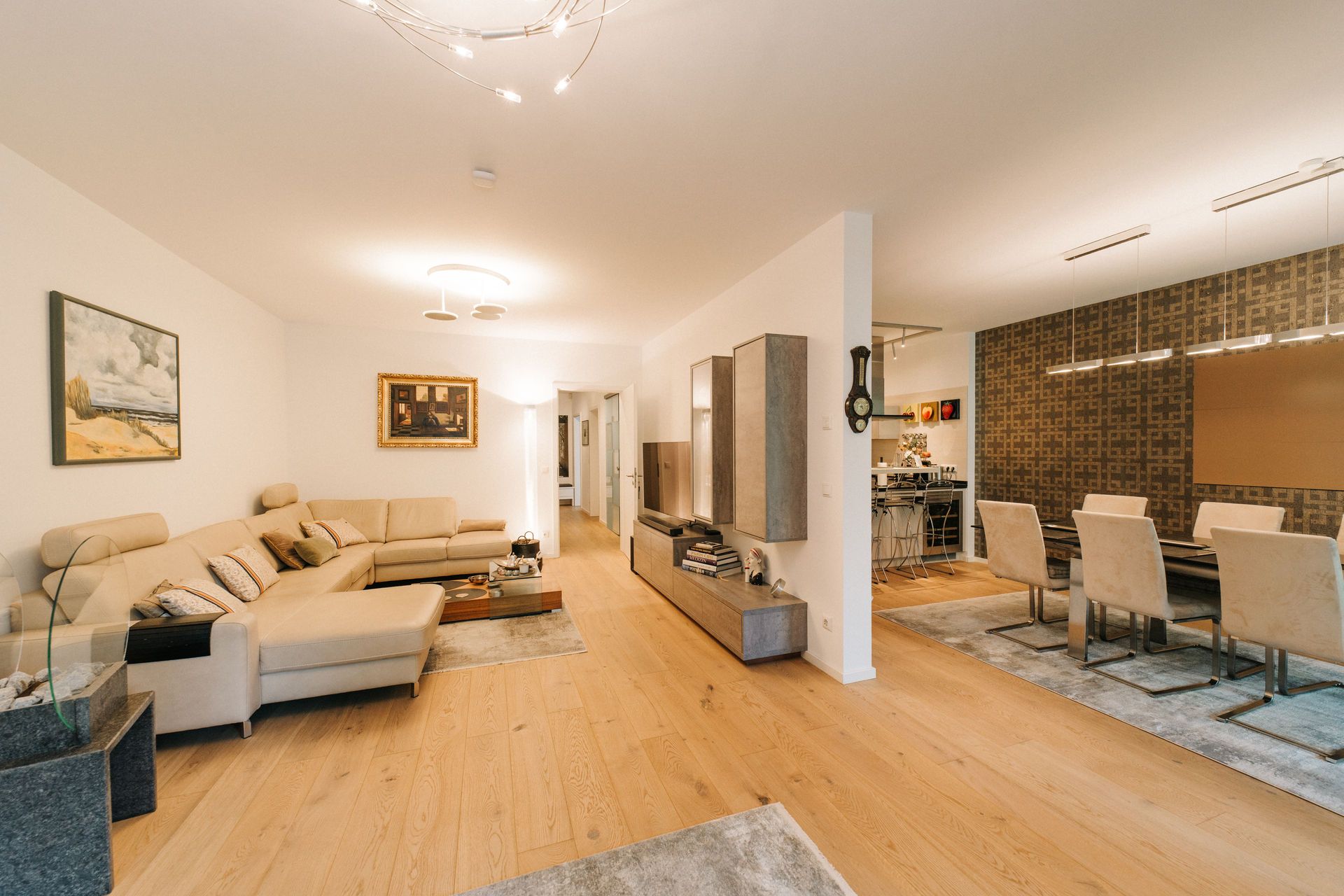 Modernes Wohnzimmer mit beigem Sofa, TV-Wand und angrenzendem Essbereich mit sechs Stühlen.