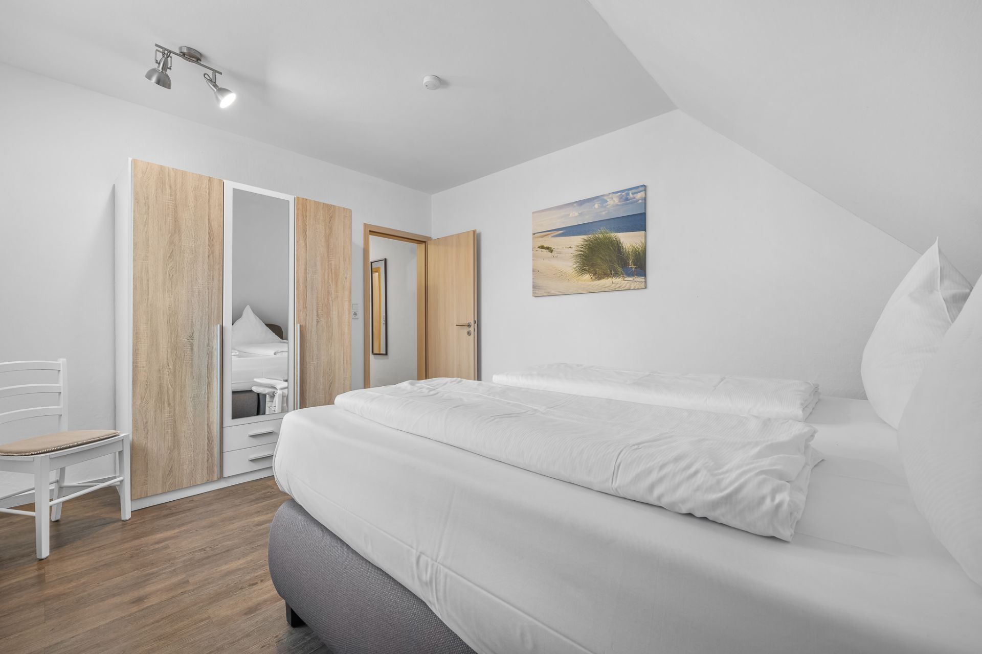 Helles Schlafzimmer mit Doppelbett, Kleiderschrank, Stuhl und Bild an der Wand.