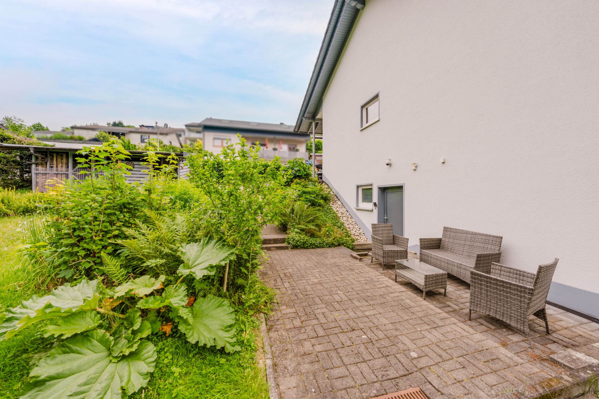 Gepflasterte Terrasse mit Gartenmöbeln neben einem weißen Wohnhaus, umgeben von grünen Pflanzen.