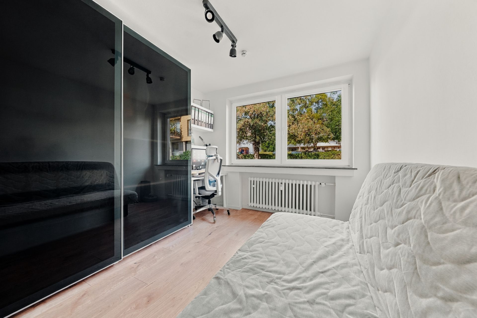 Helles Zimmer mit Schreibtisch, Stuhl, Sofa und großem schwarzen Kleiderschrank vor Fenster.