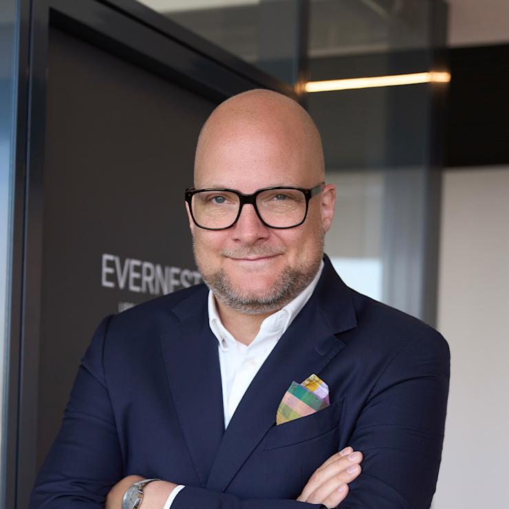 Ein Mann mit Brille und Anzug steht mit verschränkten Armen vor einer Tür.