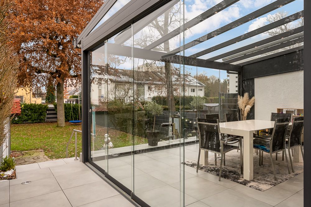 Veranda mit Glaswänden und Esstisch, Blick auf Garten und Bäume.