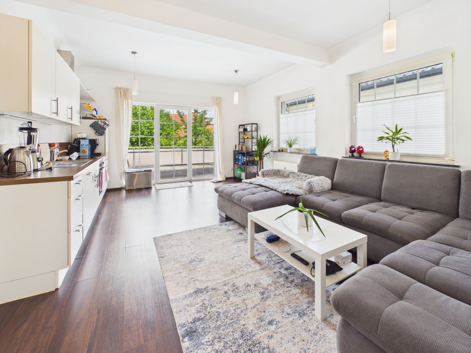 Helles Wohnzimmer mit Ecksofa, Teppich, Couchtisch und offener Küche, große Fenster und Balkonzugang.