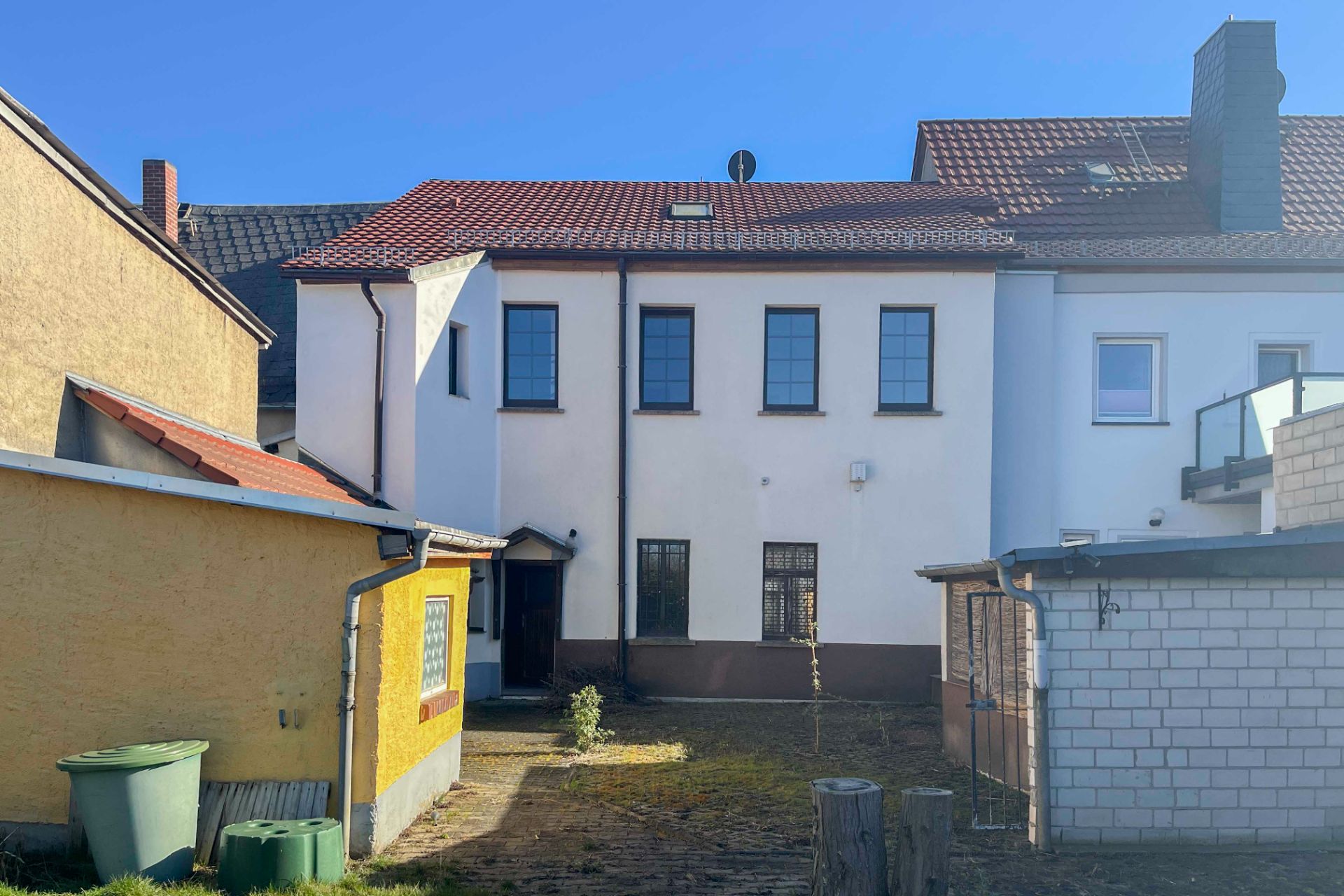 Hinterhof mit weißem Wohnhaus, Nebengebäuden und grünen Mülltonnen bei Tageslicht.
