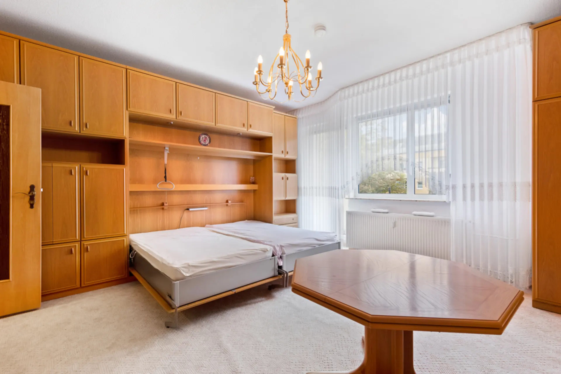 Helles Schlafzimmer mit Doppelbett, Holzmöbeln, großem Fenster und Deckenleuchter.