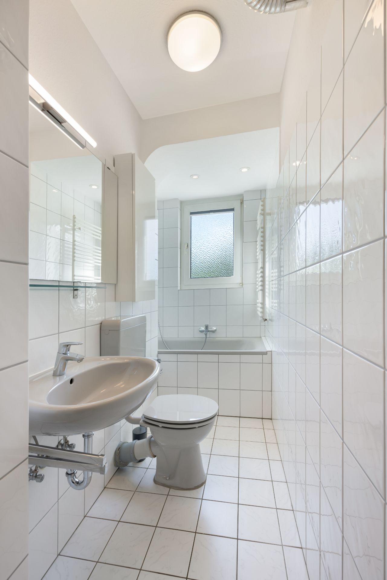 Helles Badezimmer mit Fliesen, Waschbecken, Toilette, Badewanne und Fenster.