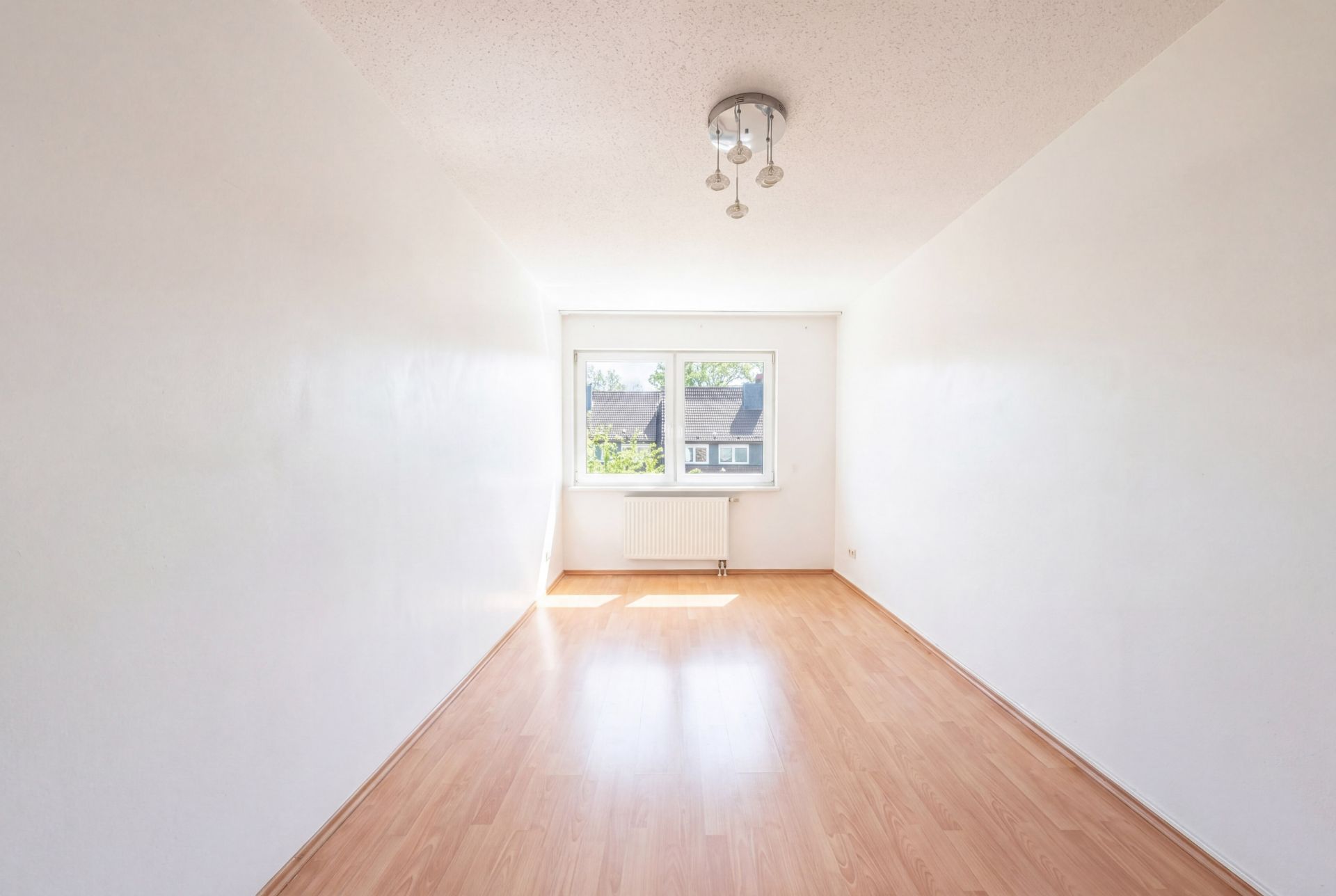 Leerer, heller Raum mit Holzfußboden, weiß gestrichenen Wänden und einem Fenster an der Stirnseite.
