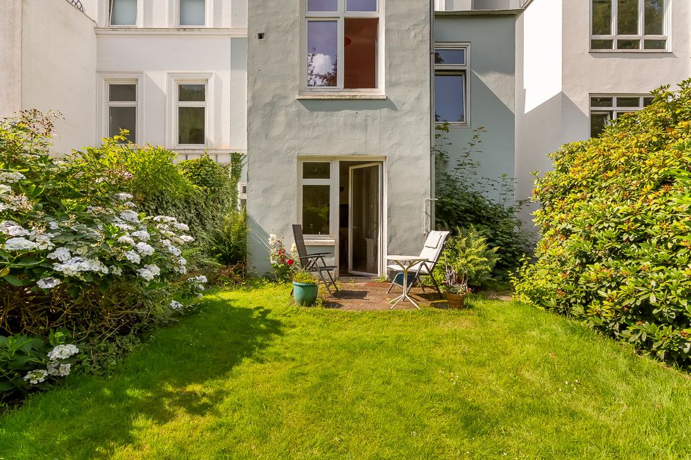 Kleiner Garten mit Terrasse, zwei Stühlen, Tisch und Pflanzen vor einem Wohnhaus.