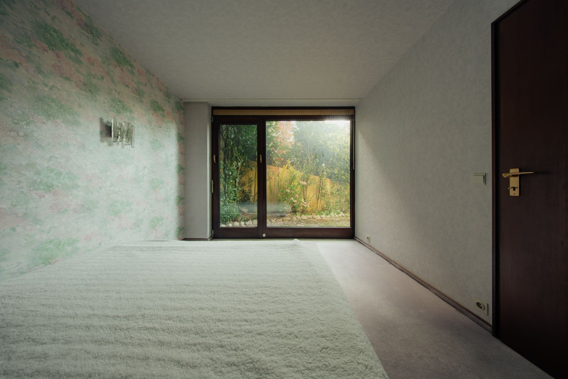 Leeres Zimmer mit Teppichboden, bunter Tapete und Glastür zum Garten.