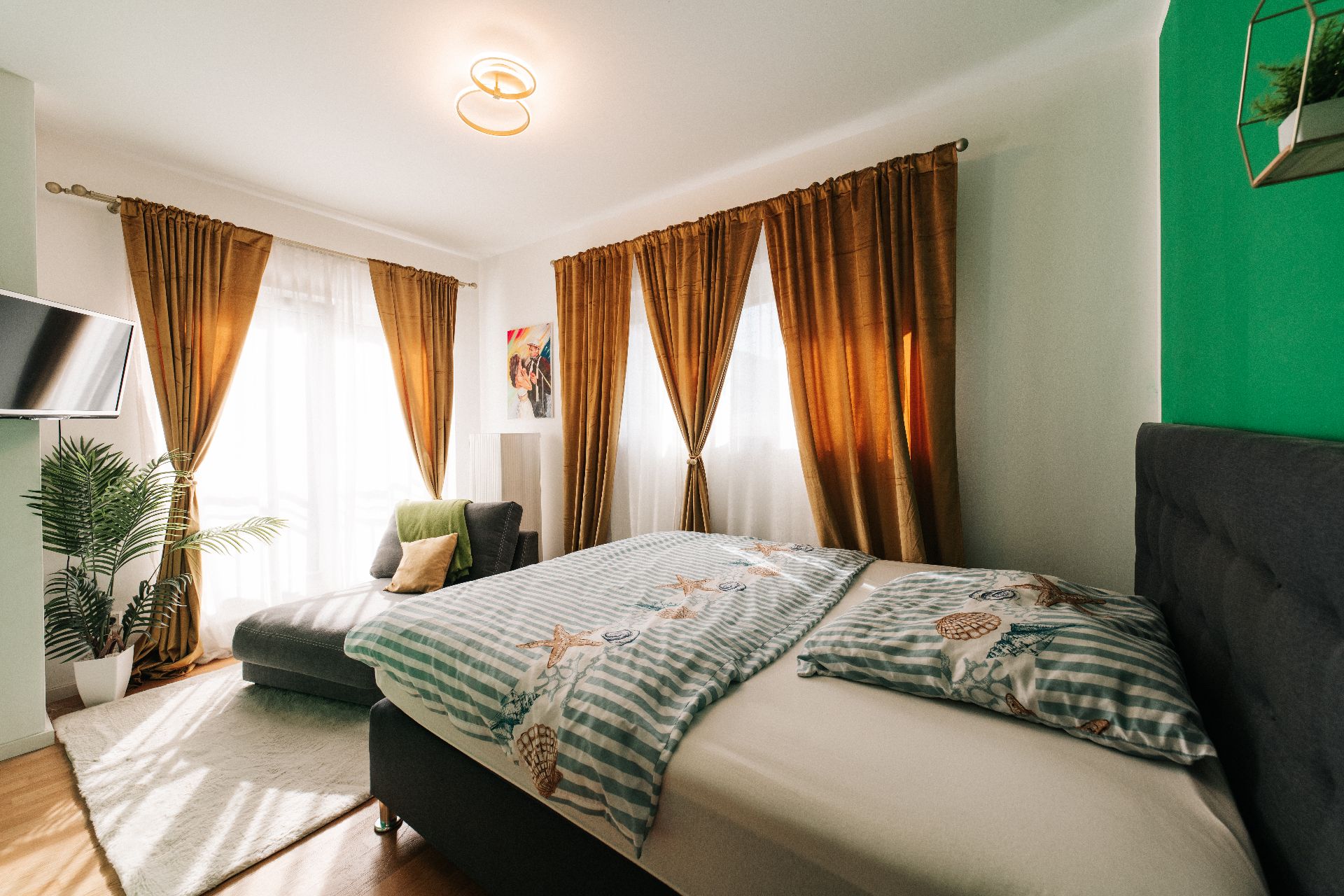 Helles Schlafzimmer mit Doppelbett, braunen Vorhängen, Sofa, Zimmerpflanze und TV an der Wand.