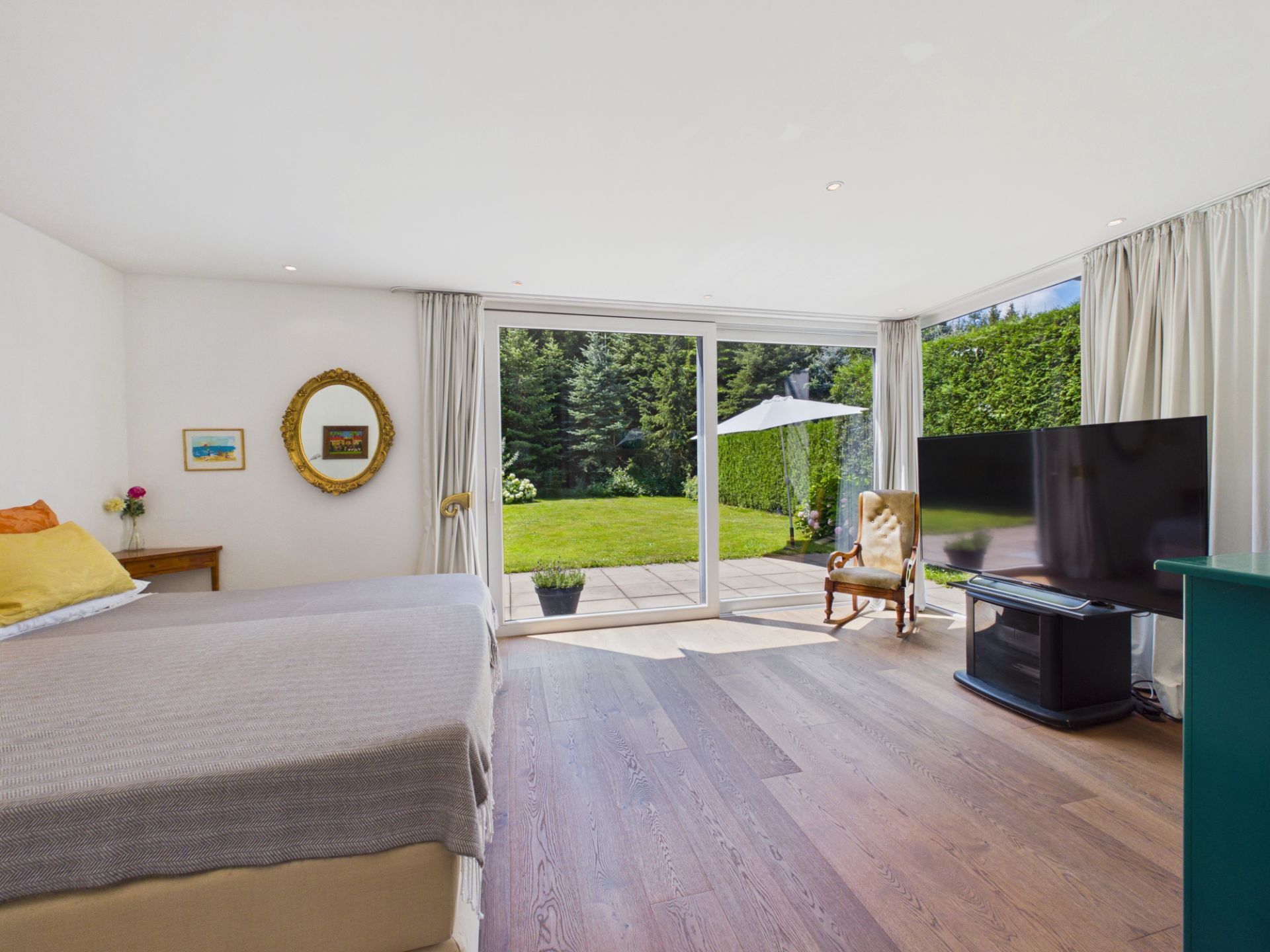 Helles Schlafzimmer mit großem Fenster, Blick auf Garten, Bett, Sessel und Fernseher.