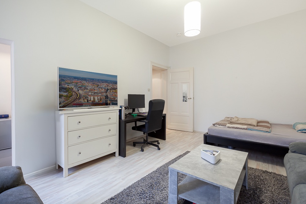 Helles Schlafzimmer mit Bett, Schreibtisch, Fernseher auf Kommode und Couchtisch auf Teppich.