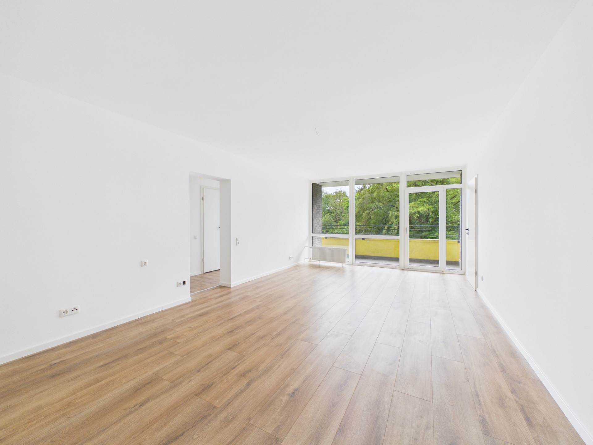 Helles, leeres Wohnzimmer mit Holzfußboden und großen Fenstern zu einem Balkon mit Blick ins Grüne.
