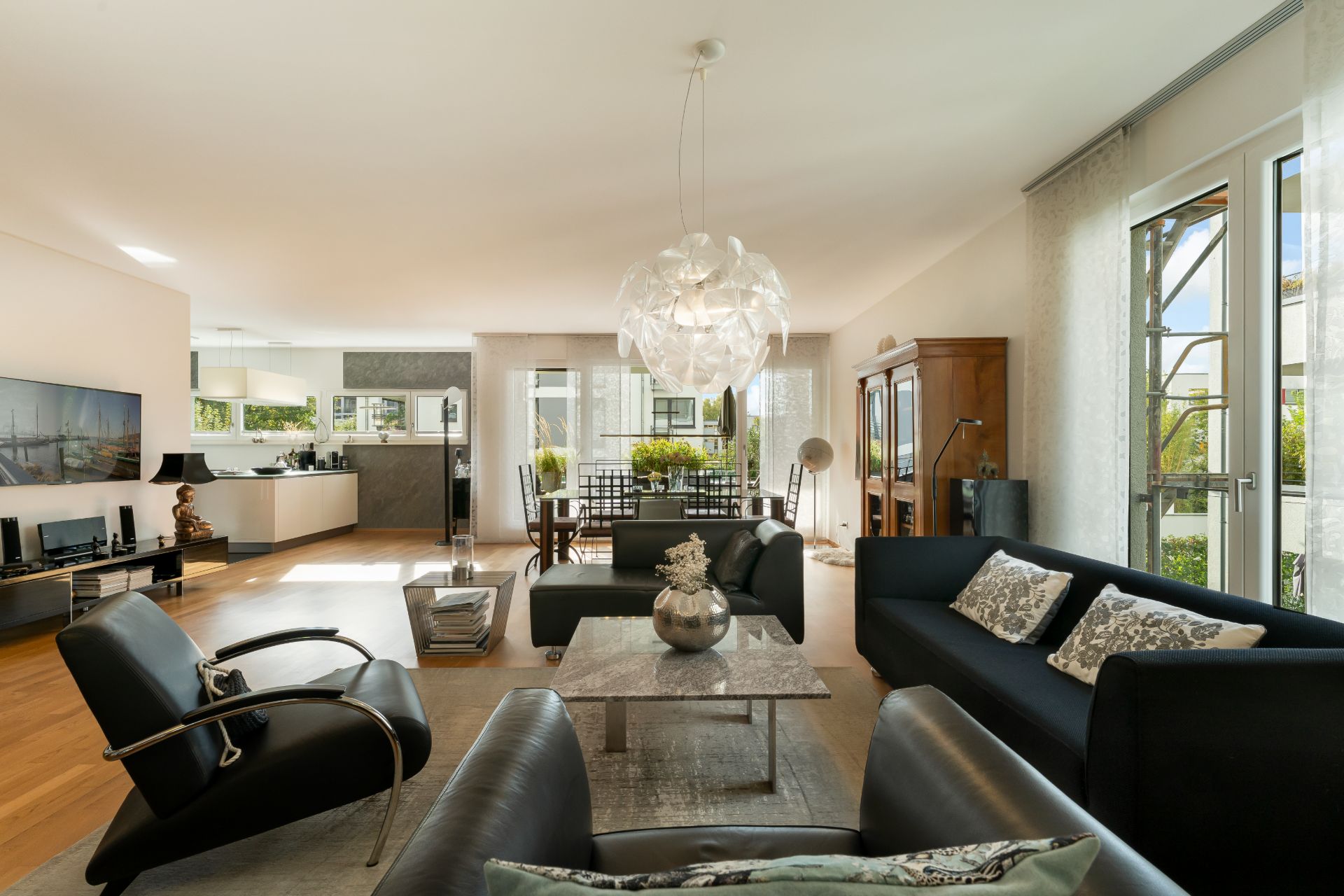 Modernes Wohnzimmer mit schwarzen Sofas, Couchtisch, großem Fenster und offener Küche im Hintergrund.