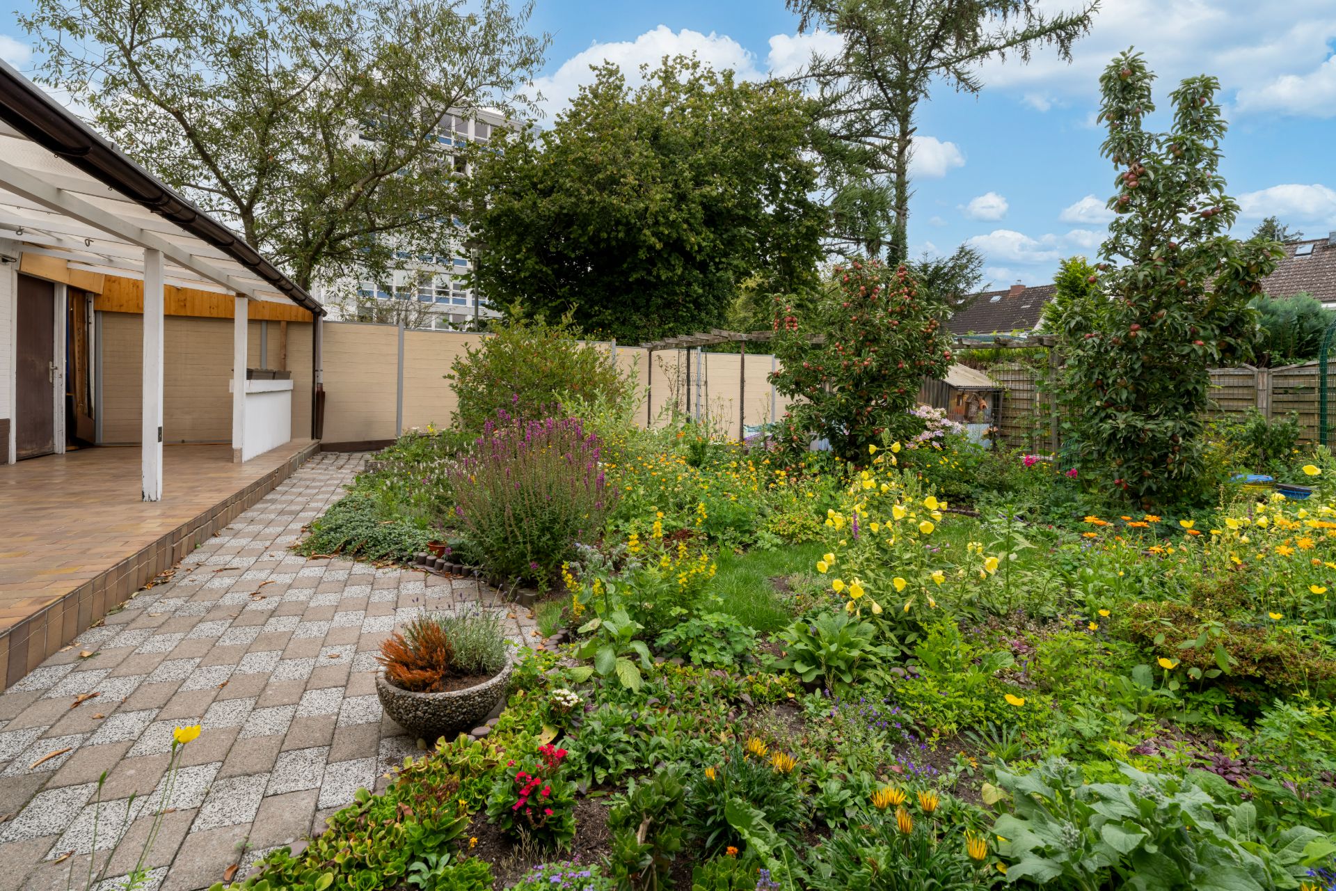 Garten mit Blumen und Pflanzen, daneben eine überdachte Terrasse mit Fliesenboden.