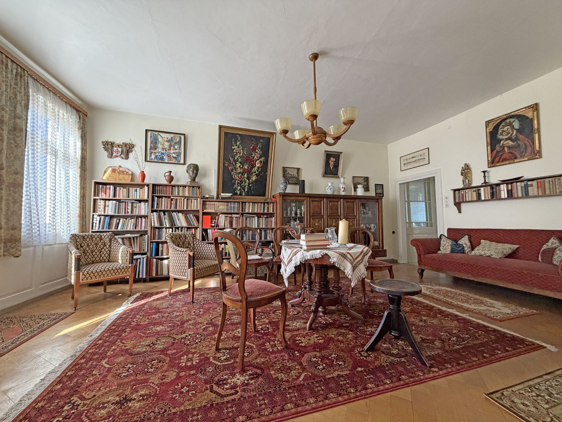 Wohnzimmer mit Teppich, Holztisch, Stühlen, Bücherregalen und rotem Sofa.
