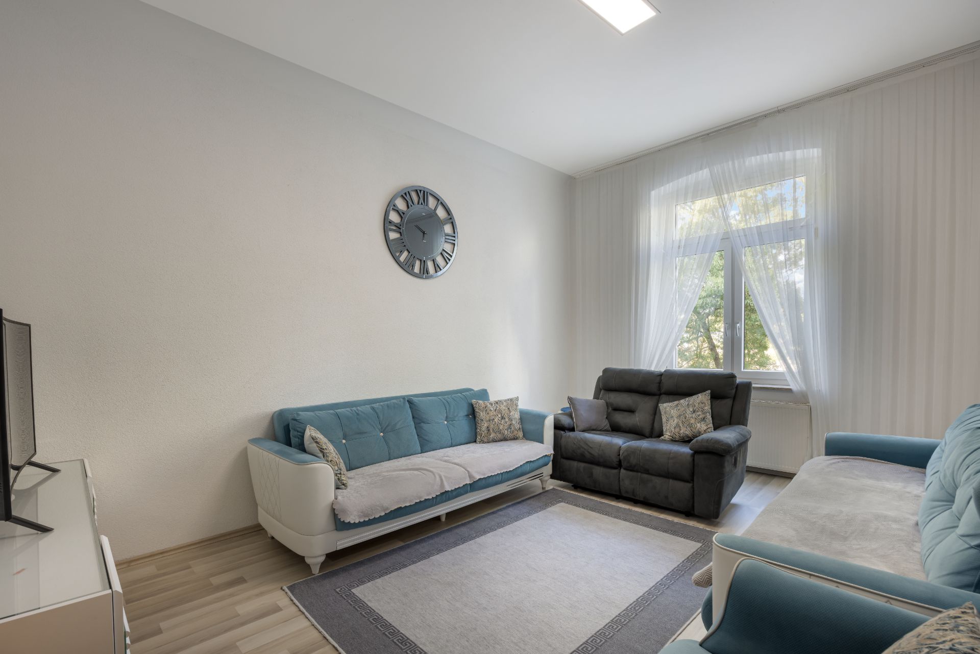 Helles Wohnzimmer mit drei Sofas, einem Teppich, einer Wanduhr und einem Fenster mit Vorhängen.