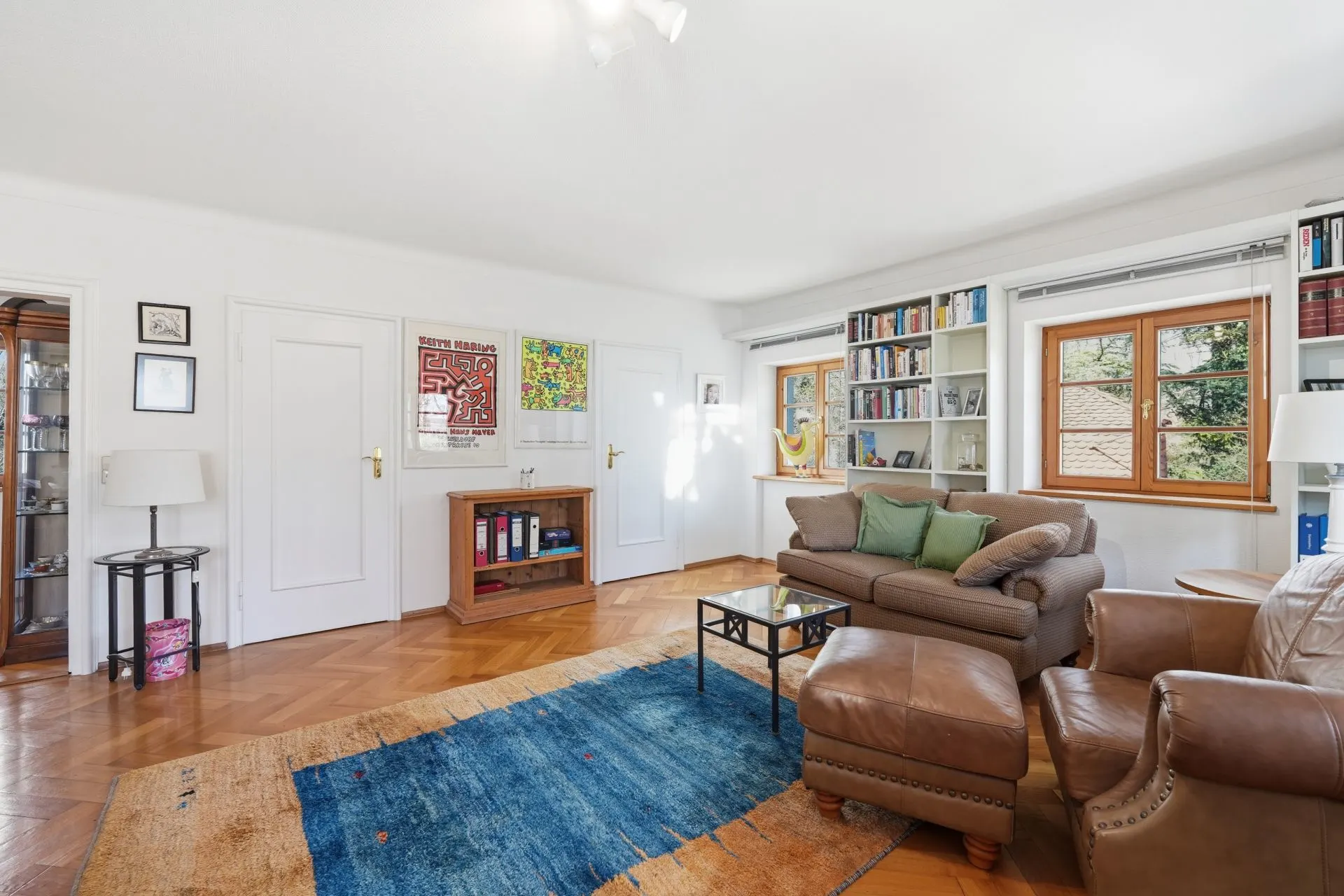 Helles Wohnzimmer mit Ledersofa, Bücherregal, Teppich und zwei Fenstern.