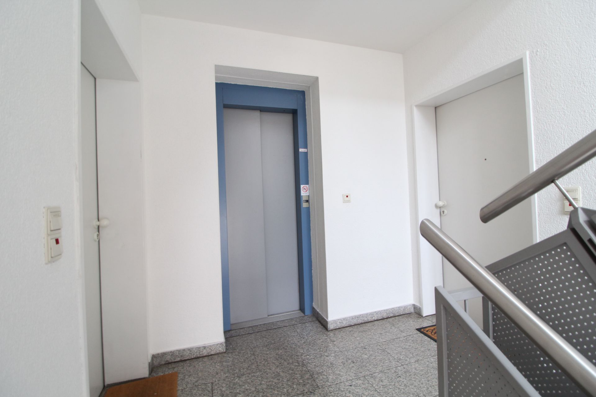 Flur mit Aufzug, zwei weißen Türen und Treppengeländer in einem Wohngebäude.