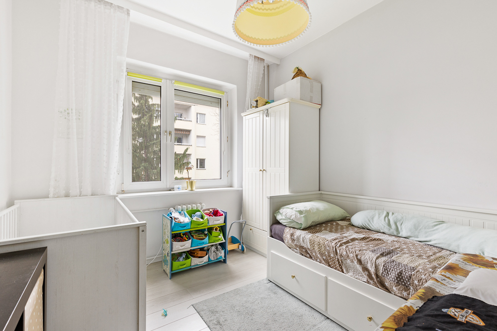 Helles Kinderzimmer mit Bett, Babybett, Kleiderschrank und Spielzeugregal vor einem Fenster.