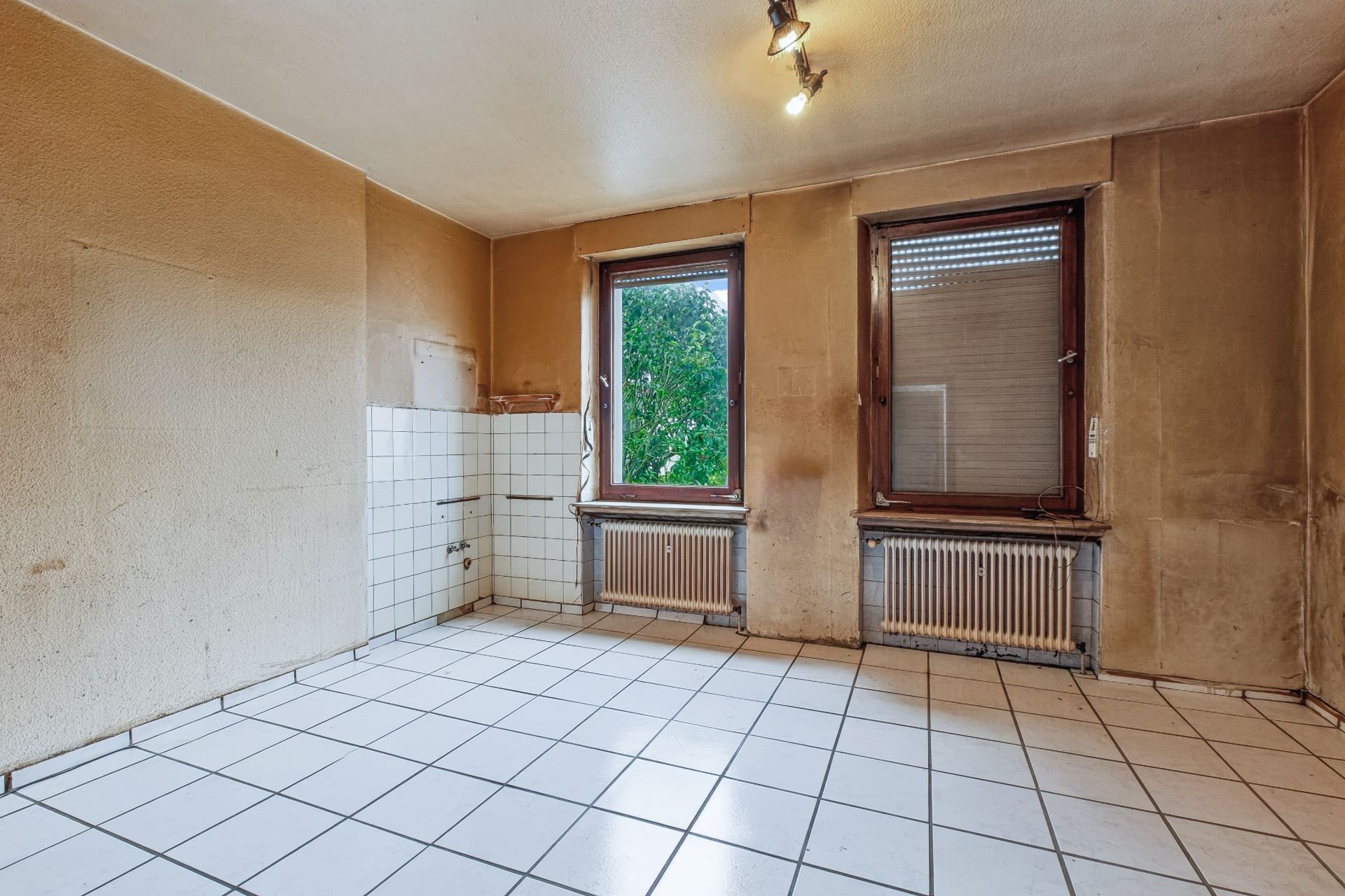 Leerer Raum mit weißen Fliesen, zwei Fenstern, Heizkörpern und gefliester Ecke.