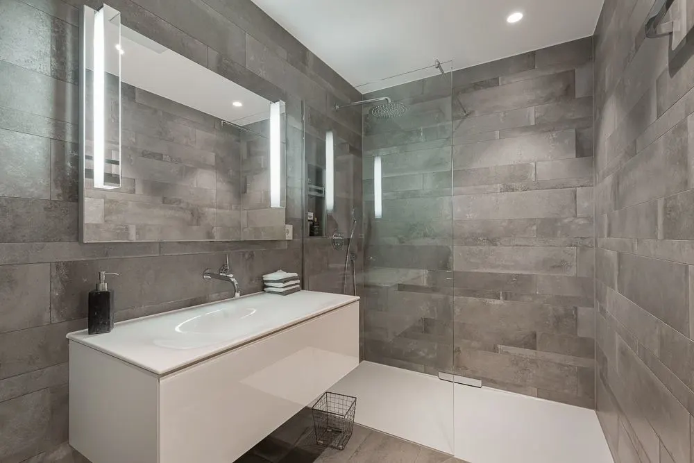 Modernes Bad mit großem Spiegel, Waschbecken, Dusche mit Glaswand und grauen Fliesen.