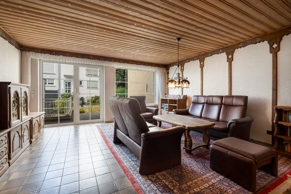 Wohnzimmer mit braunen Ledermöbeln, Holztisch, Teppich und großen Fenstern zur Terrasse.