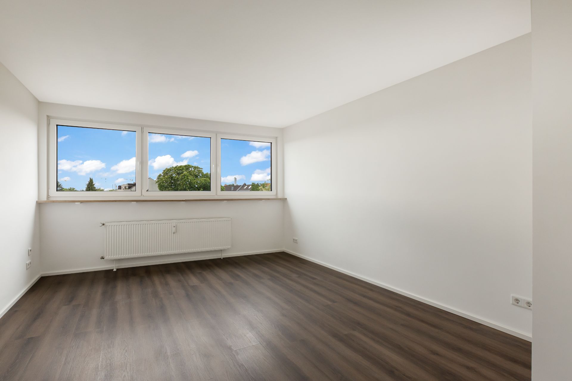 Helles, leeres Zimmer mit drei großen Fenstern und dunklem Holzboden.