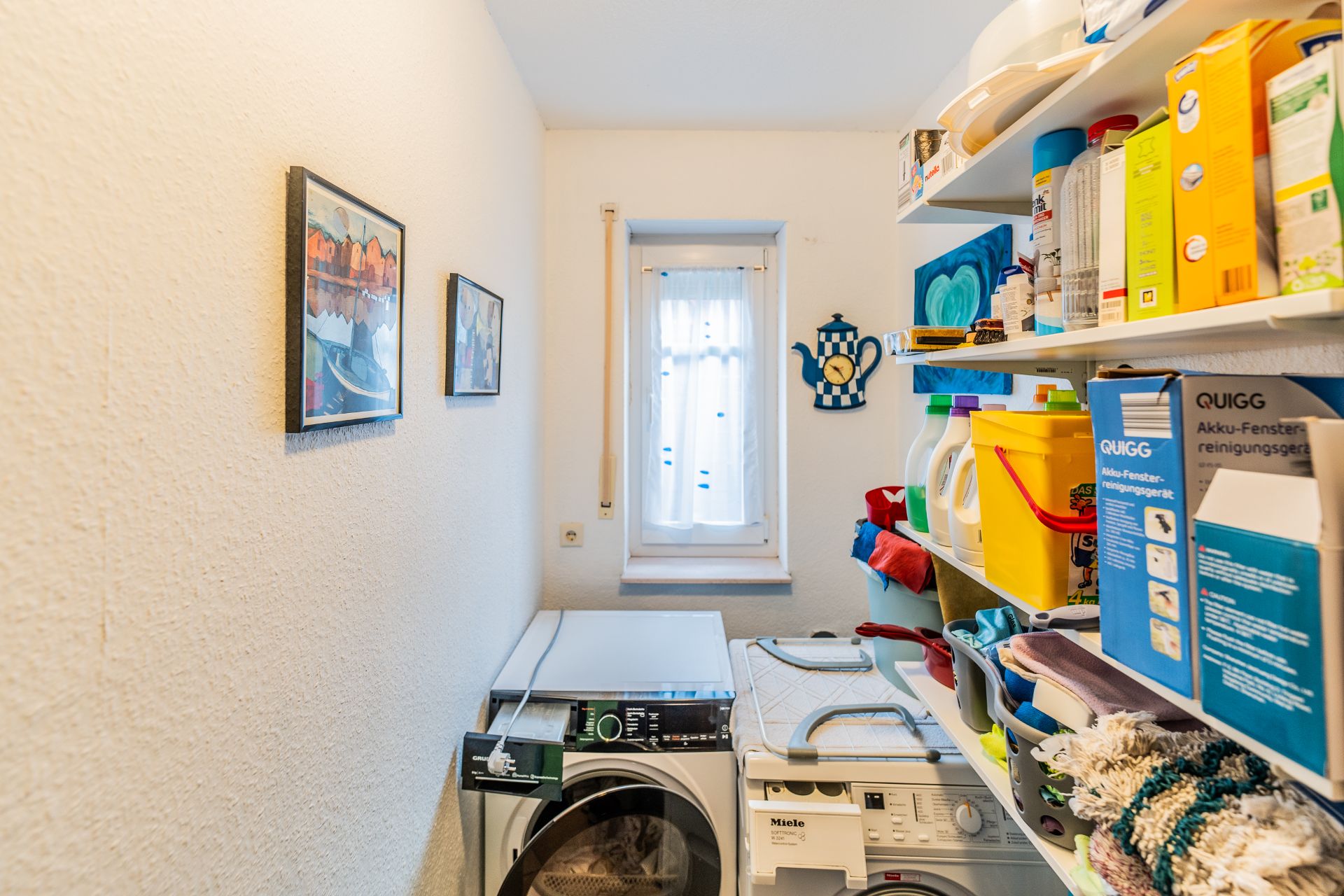Kleiner Hauswirtschaftsraum mit Waschmaschine, Trockner, Regalen und Fenstervorhang.
