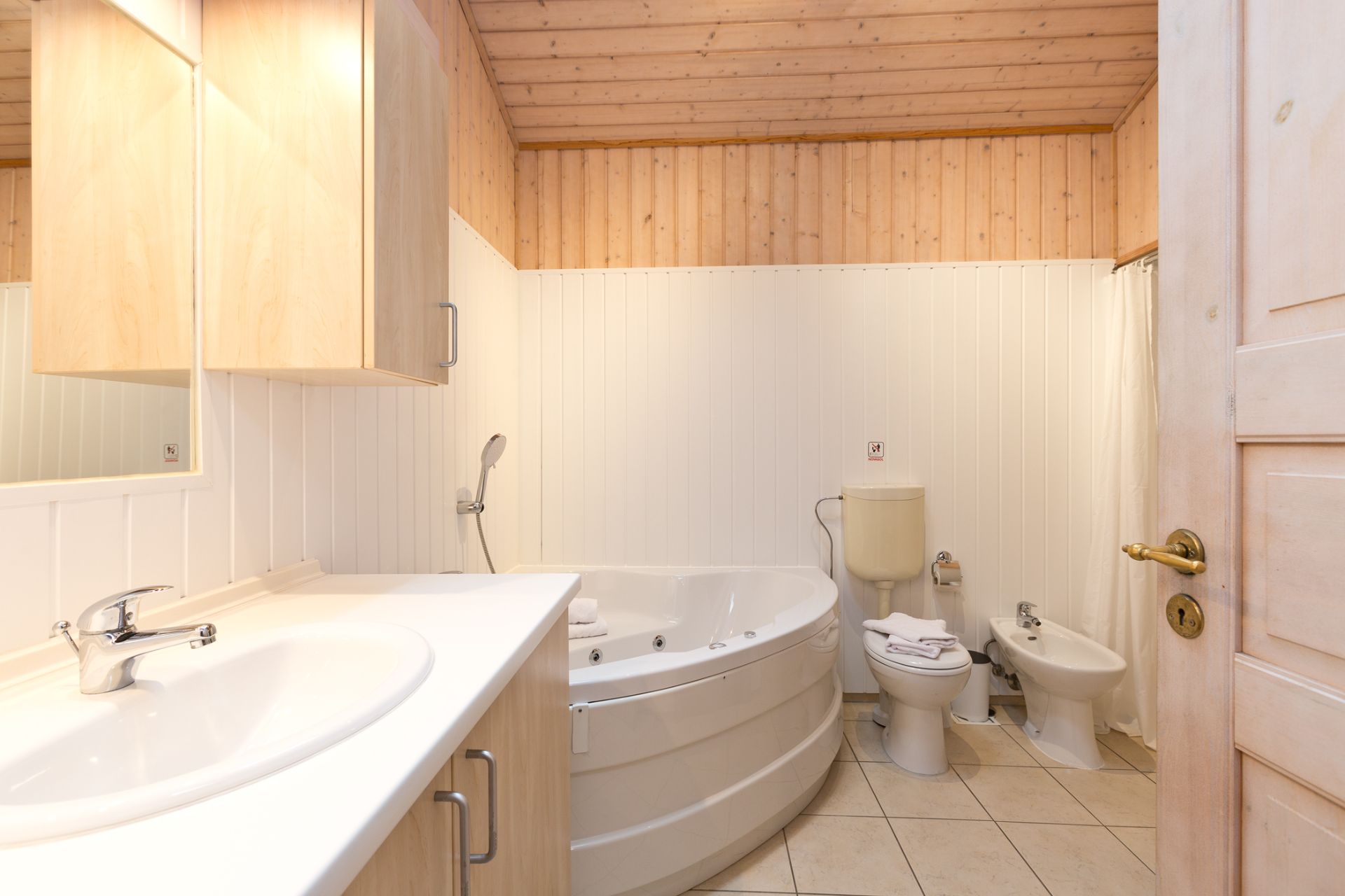 Helles Badezimmer mit Eckbadewanne, Waschbecken, Toilette und Bidet, Holzdecke und hellen Fliesen.