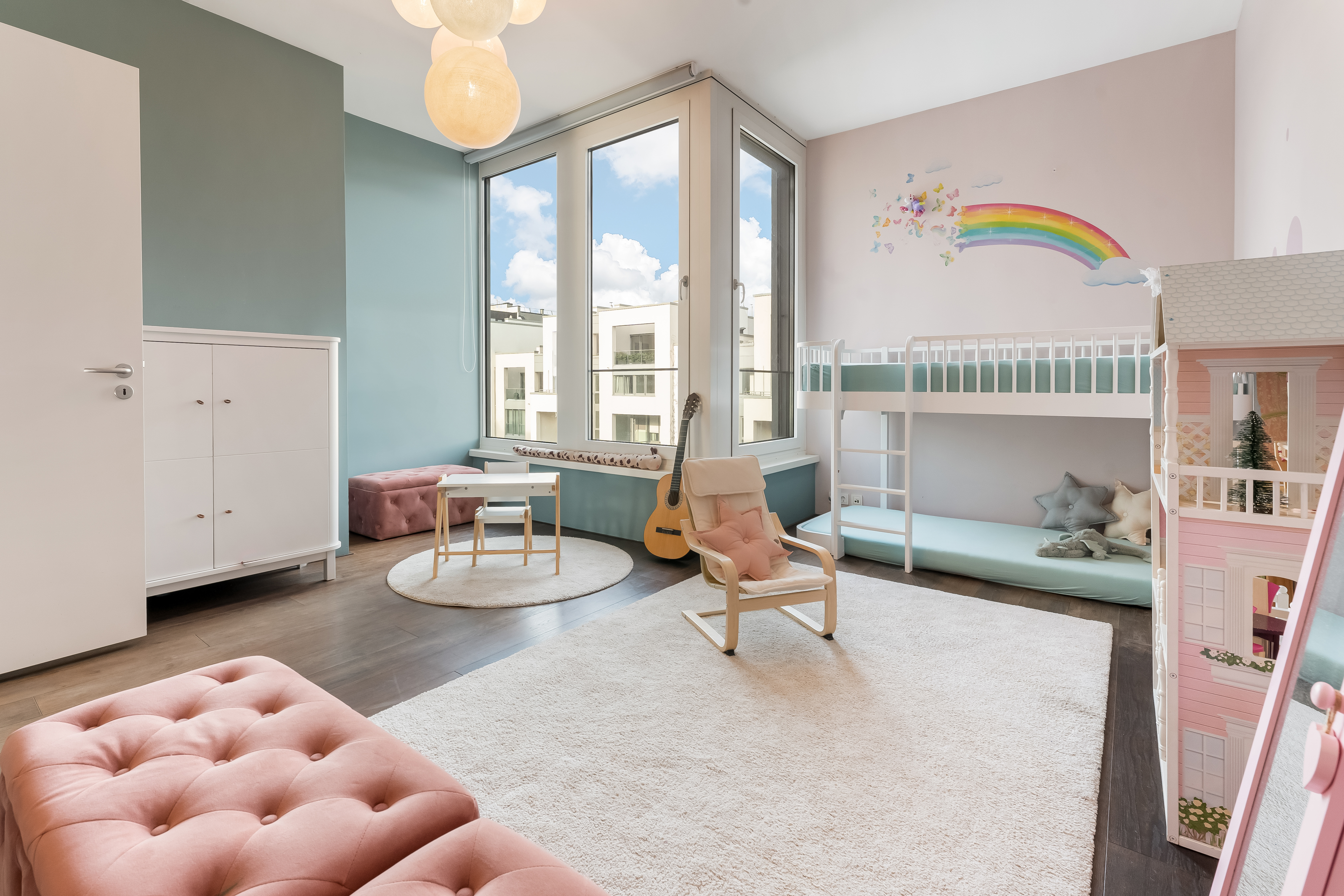 Helles Kinderzimmer mit Hochbett, großem Fenster, Teppich und Wandbild eines Regenbogens.