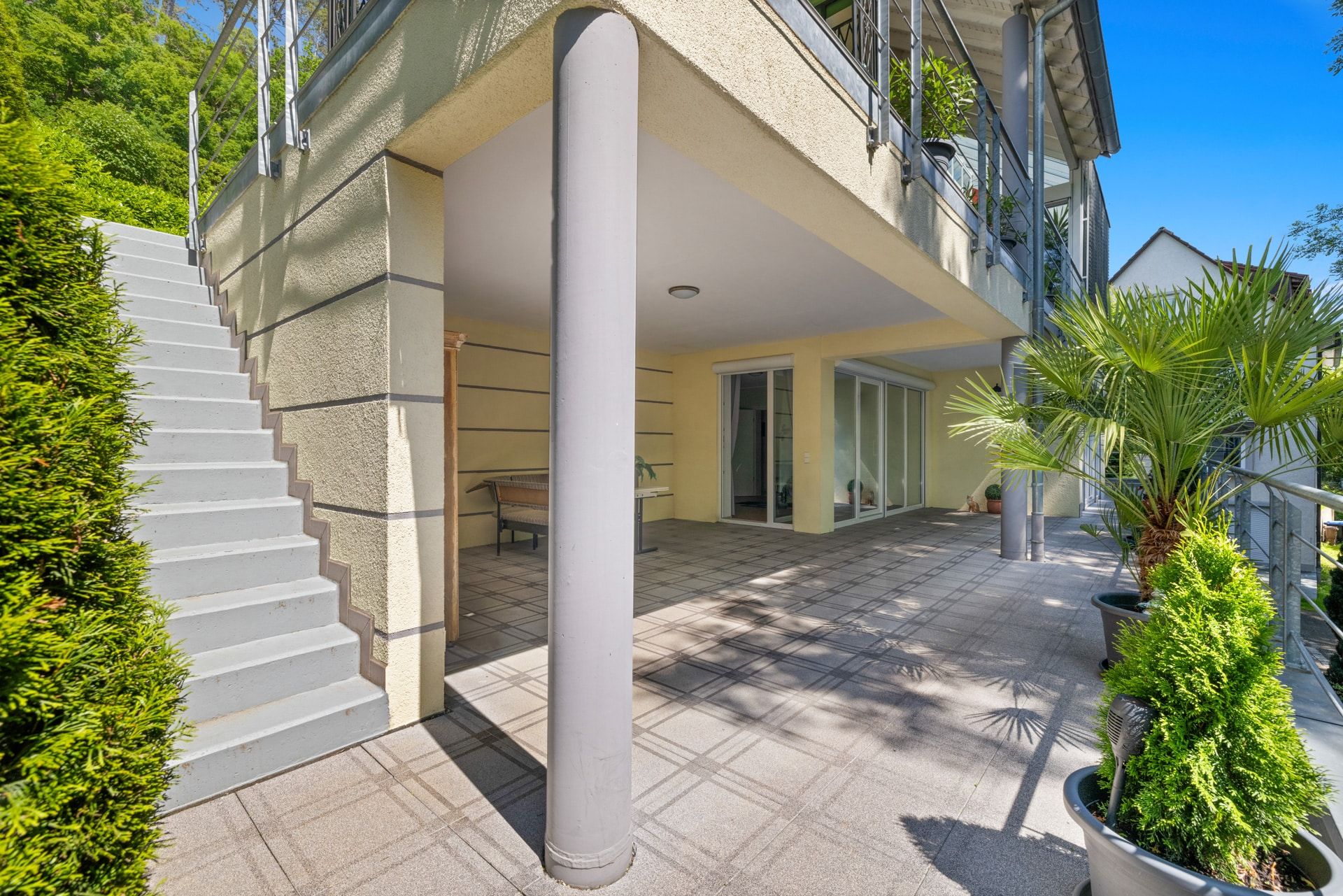 Große überdachte Terrasse mit Pflanzen, Treppe nach oben und Zugang zu Wohnräumen durch Glasschiebetüren.