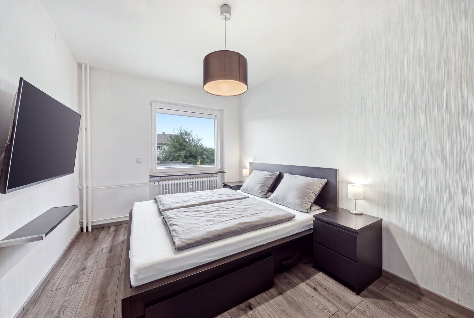 Helles Schlafzimmer mit Doppelbett, Nachttischen, TV an der Wand und einem Fenster mit Ausblick.