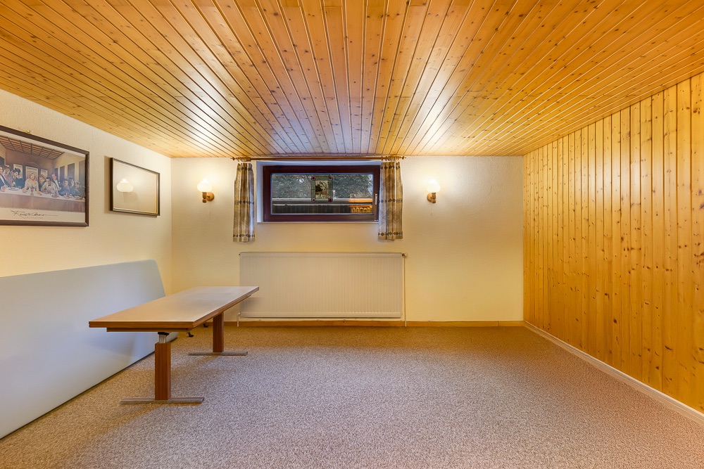 Holzvertäfelter Raum mit Tisch, Fenster mit Gardinen, Wandbildern und Wandlampen.