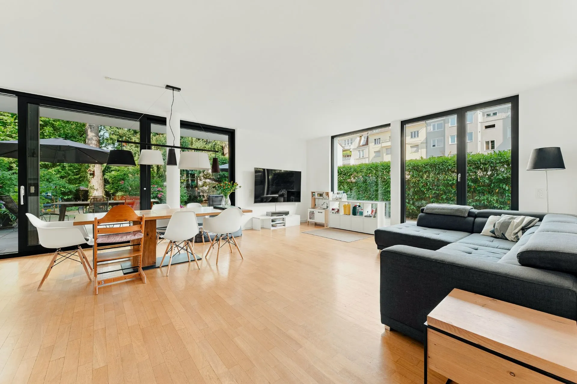 Modernes Wohnzimmer mit großer Ecksofa, Esstisch, Stühlen und großen Fenstern zum Garten.