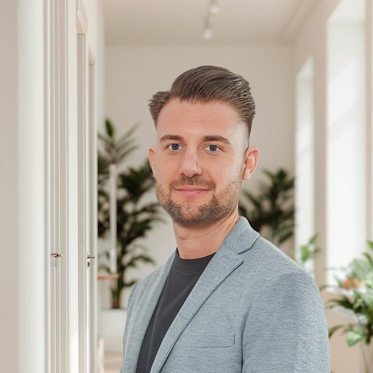 Ein Mann im grauen Sakko steht in einem hellen Raum mit Pflanzen und großen Fenstern.